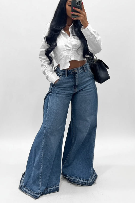Person wearing a white shirt and blue jeans standing next to a plant indoors.